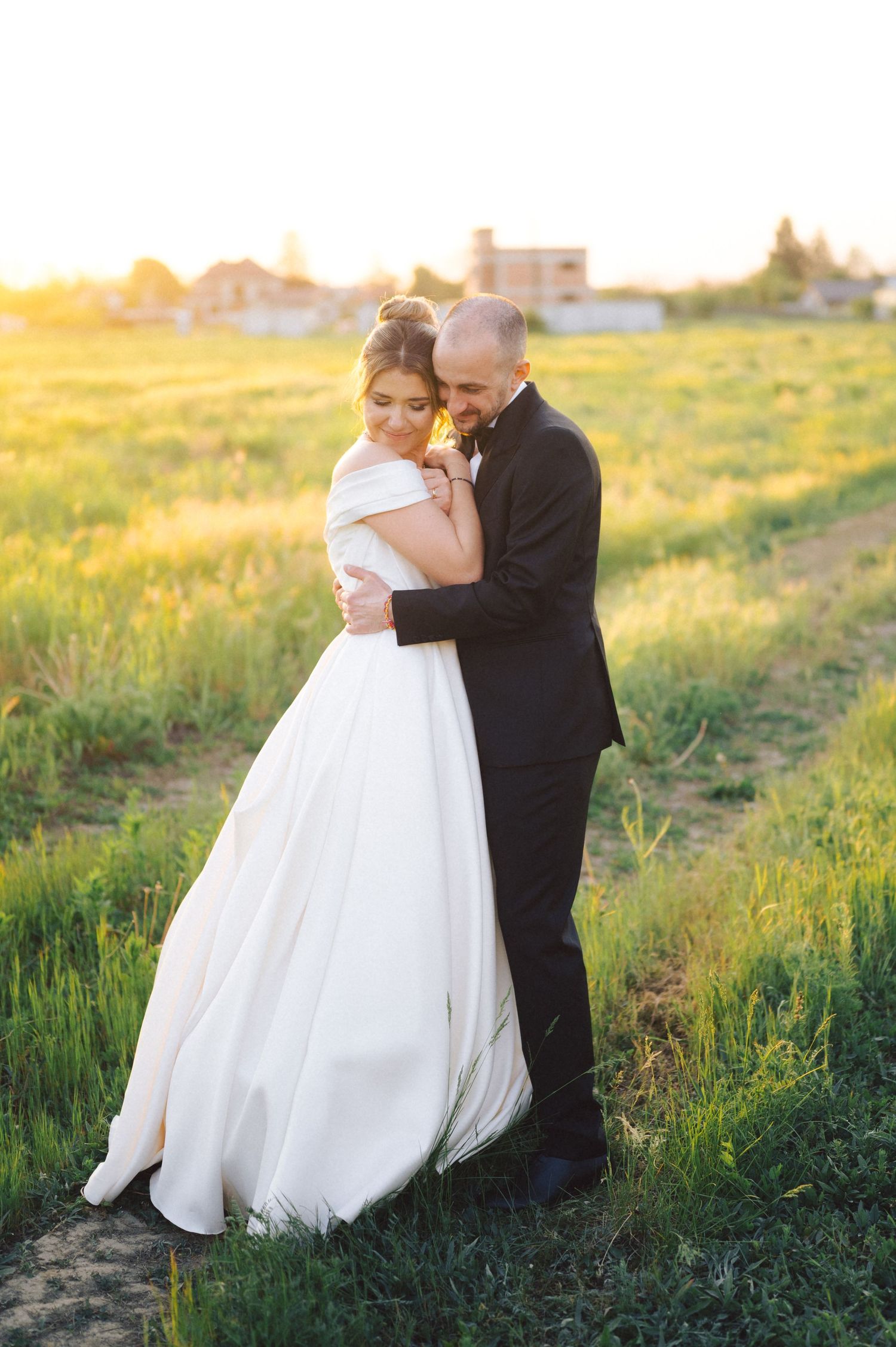 A romantic wedding couple embraces in a sunlit field during golden hour with buildings in the background.