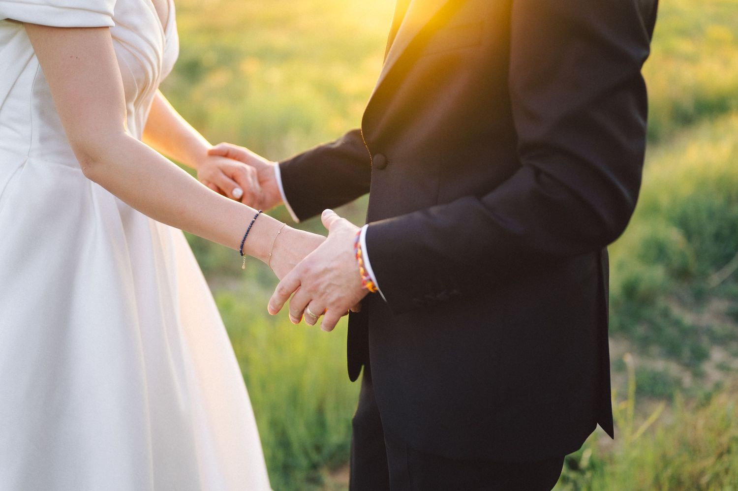 Close-up of couple holding hands in wedding attire with sunset light glowing through grass in background.