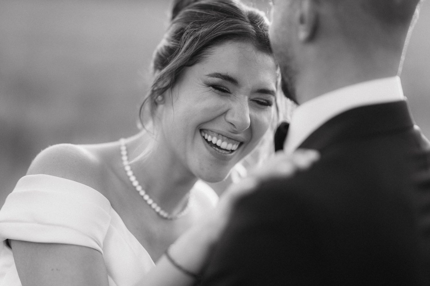 Black and white wedding portrait of bride wearing pearl necklace smiling joyfully at groom in intimate moment.