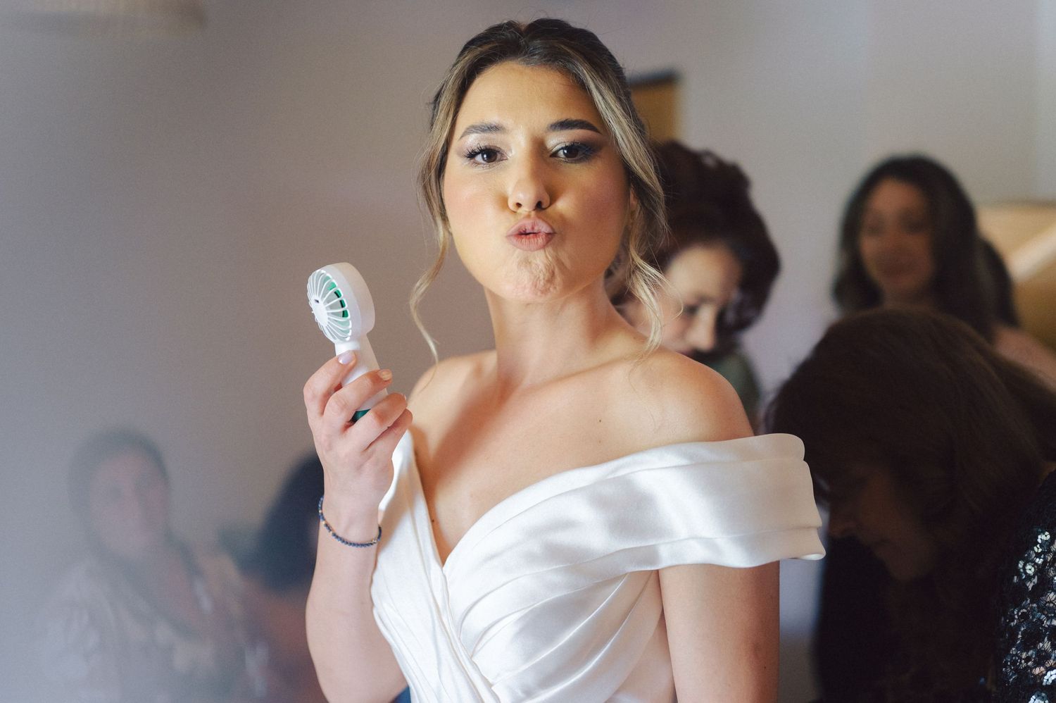 Someone in a white off-shoulder wedding dress makes a playful expression while holding up a message in a softly lit room.