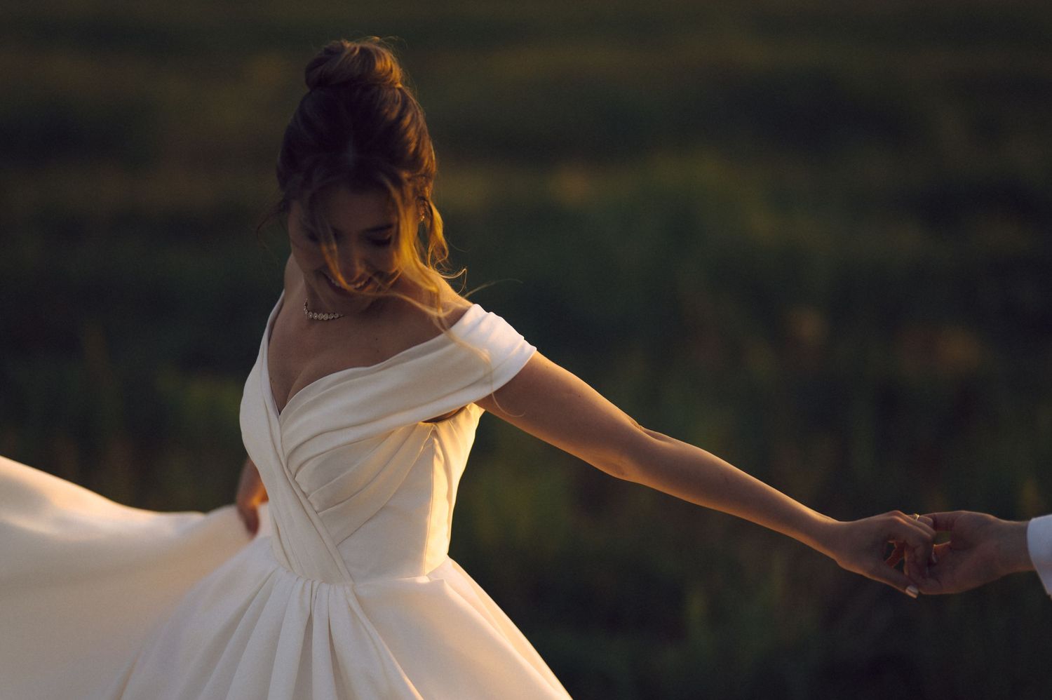 A bride in white wedding dress twirls gracefully in golden sunlight while holding someone's hand at sunset.