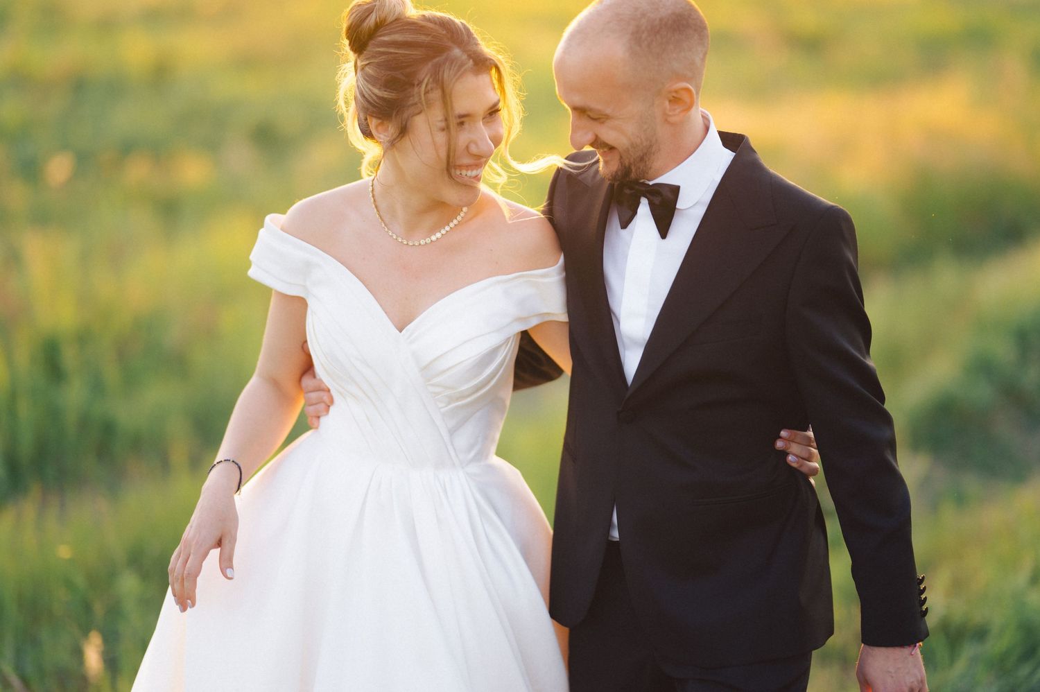 A bride in an off-shoulder white gown and groom in black tuxedo walking together through a sunlit meadow.