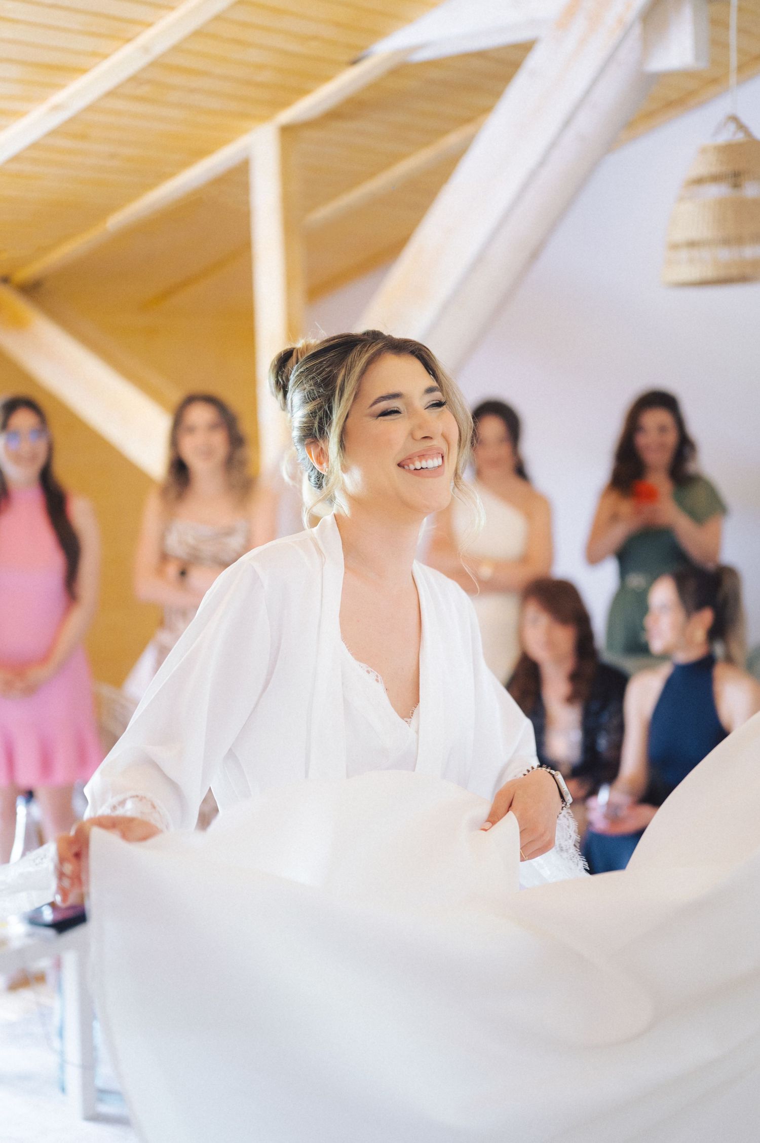 A person in a flowing white wedding dress spins joyfully while others watch in a sunlit room with wooden beams.