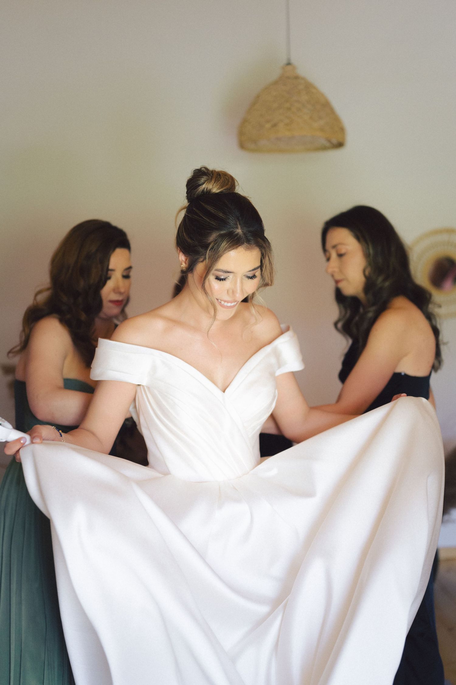 A bride in an off-shoulder white wedding gown gets help from bridesmaids wearing sage green dresses while getting ready.