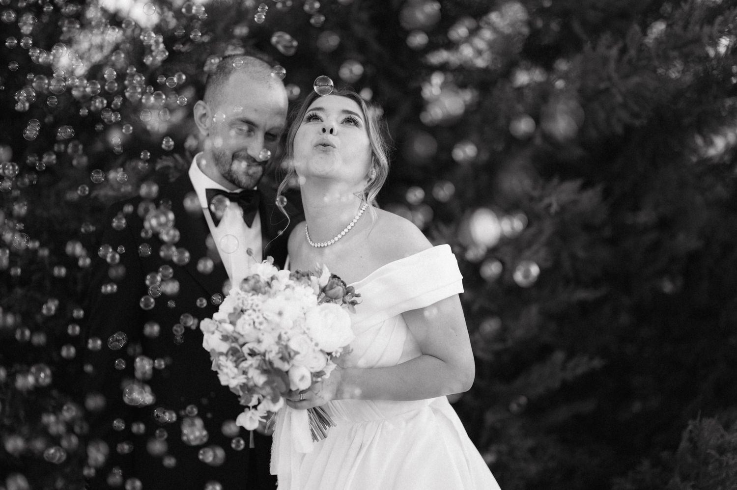 Black and white wedding photo of a couple surrounded by bubbles as the bride blows them in her white gown.