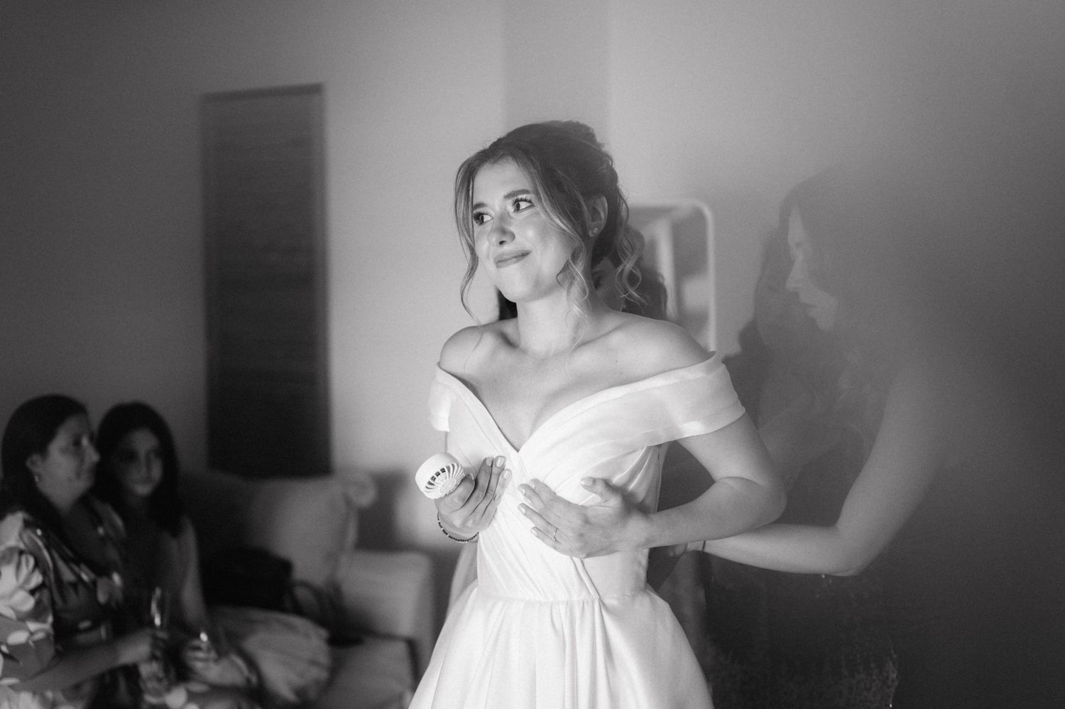 Black and white photograph showing a person wearing an elegant off-shoulder white wedding dress in an indoor setting.