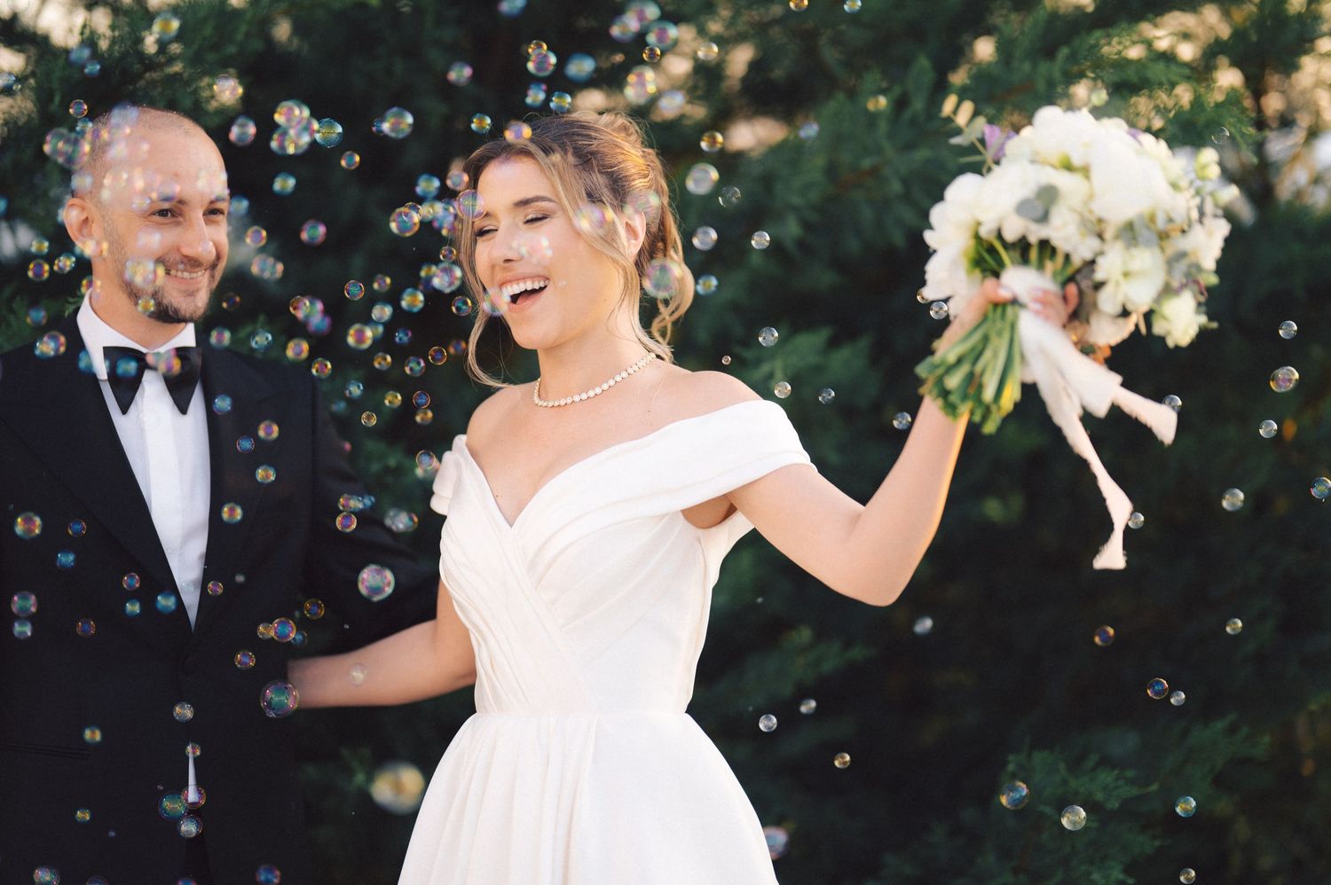 A joyful wedding moment with bubbles floating around as the bride holds a white bouquet.