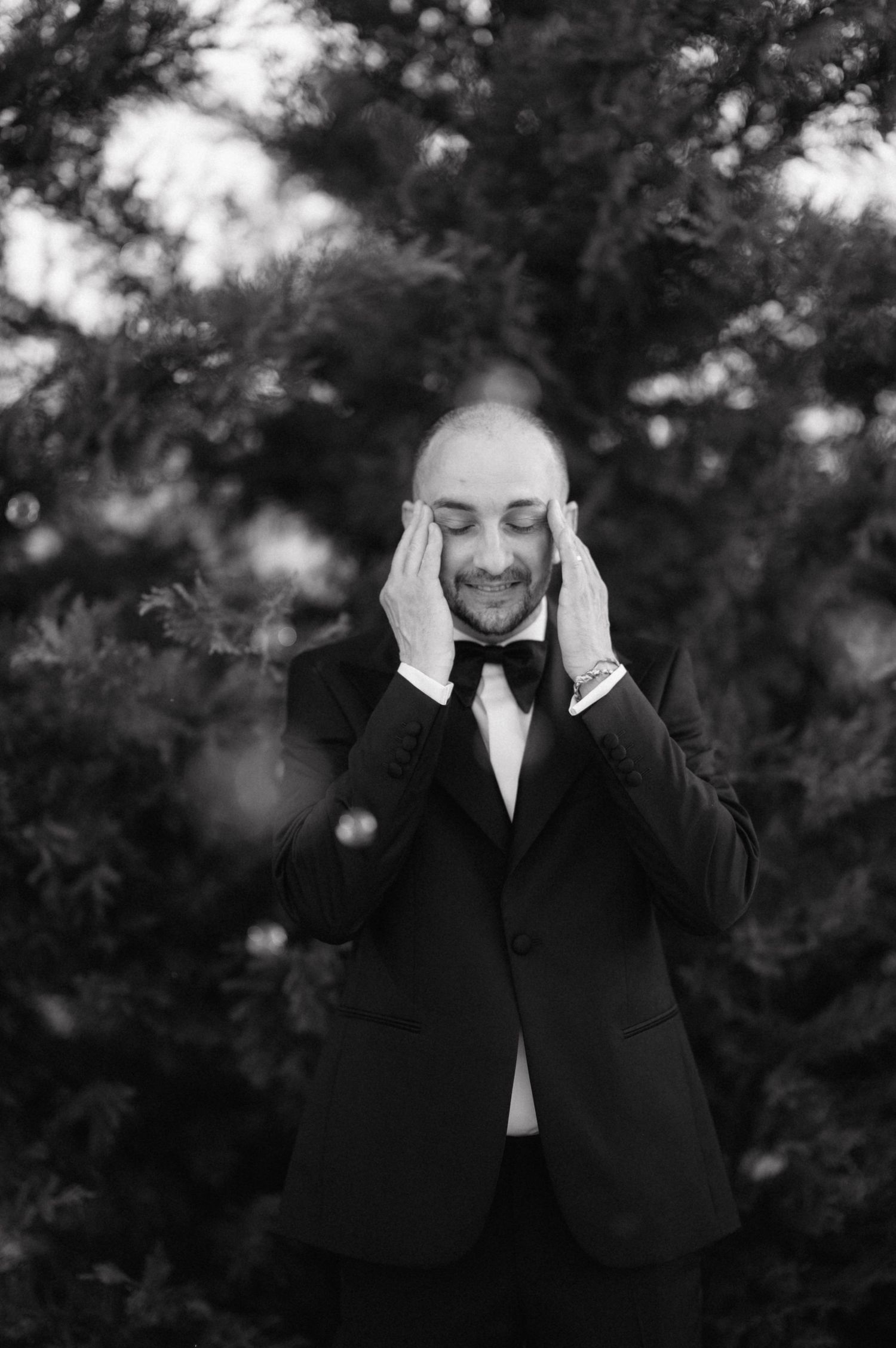A groom in a black tuxedo and bowtie holds his hands to his face with closed eyes in an emotional black and white moment.