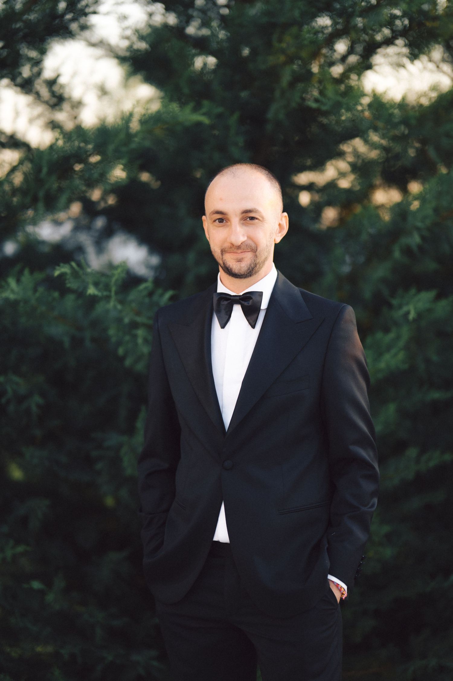 A person in a classic black tuxedo and bow tie stands outdoors with greenery in the background during golden hour.