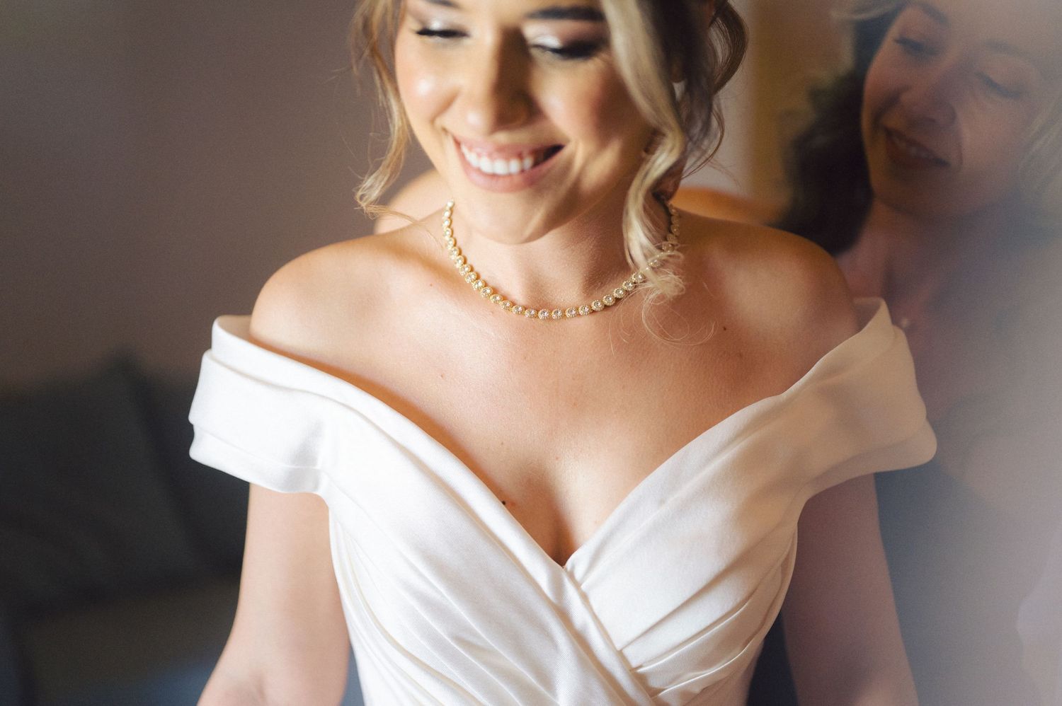 A bride in an elegant off-the-shoulder white wedding dress and delicate necklace smiles warmly.
