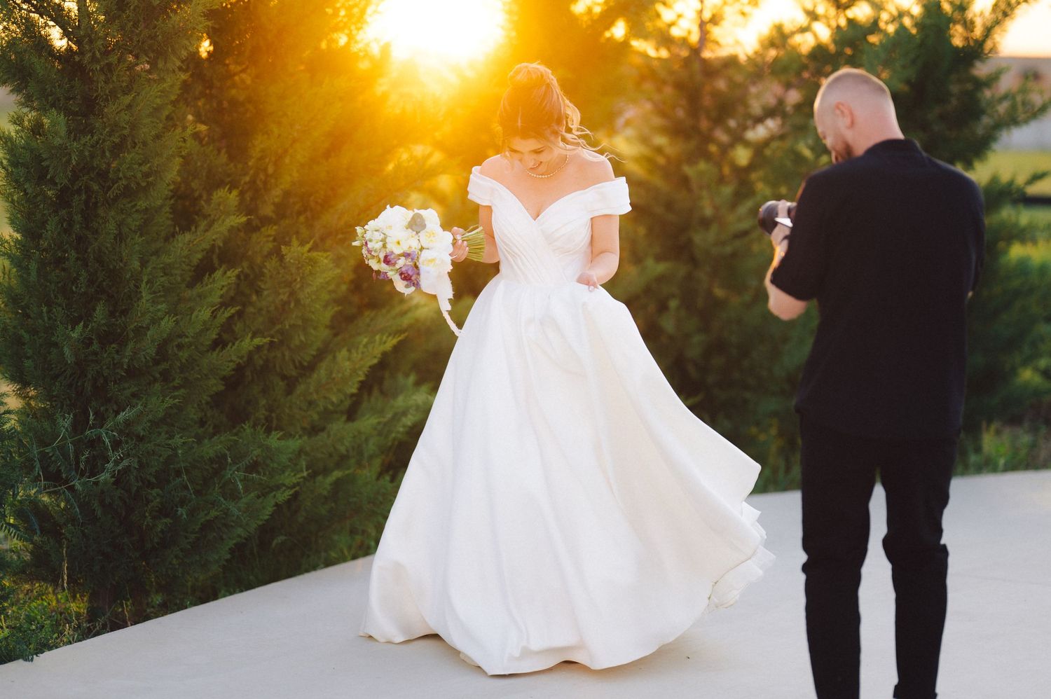 A bride in a flowing white gown walks outdoors during golden hour sunset while being photographed.