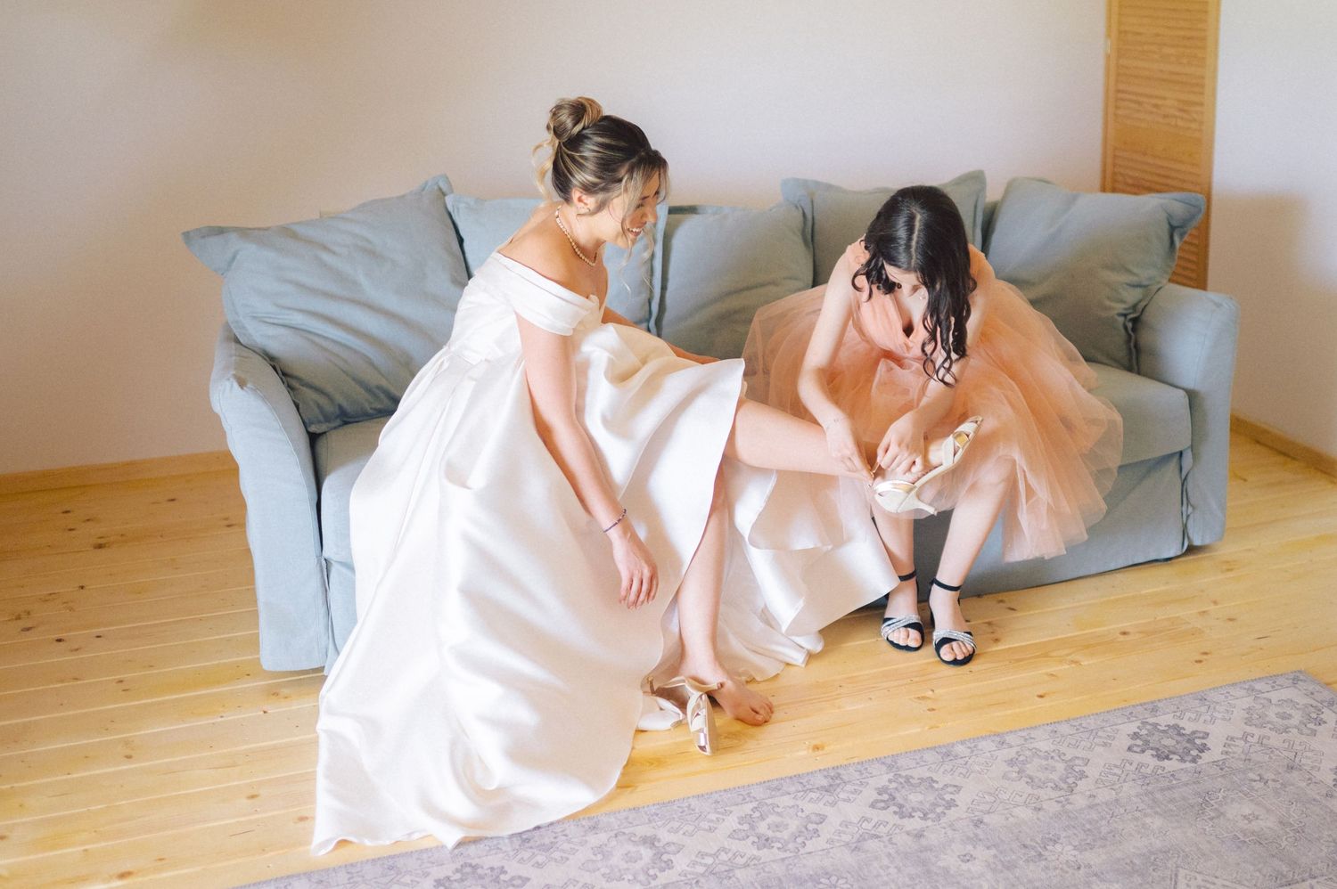 Two people in flowing white gowns sit side by side on the floor next to a gray couch in a minimalist room.