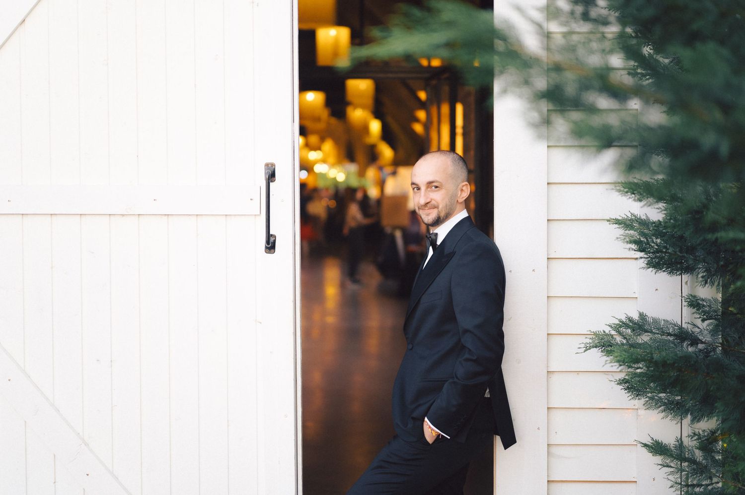 A groom in a dark suit stands casually by a white barn door with glowing lights in the background.