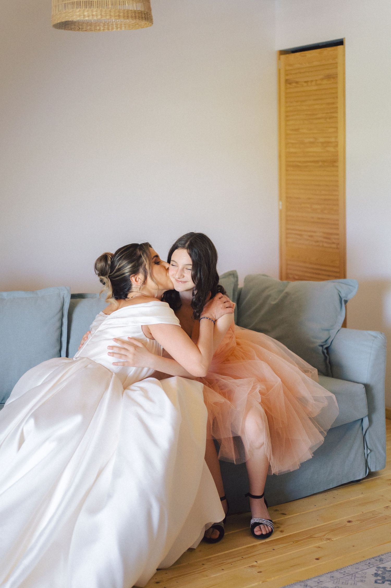 A tender moment shared between two people in formal dresses sitting on a blue couch in a sunlit room.