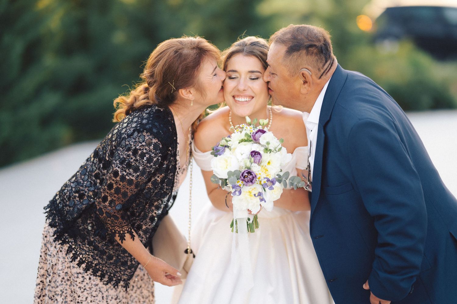 Bride in white wedding dress holding colorful bouquet receives loving kisses from family members outdoors.
