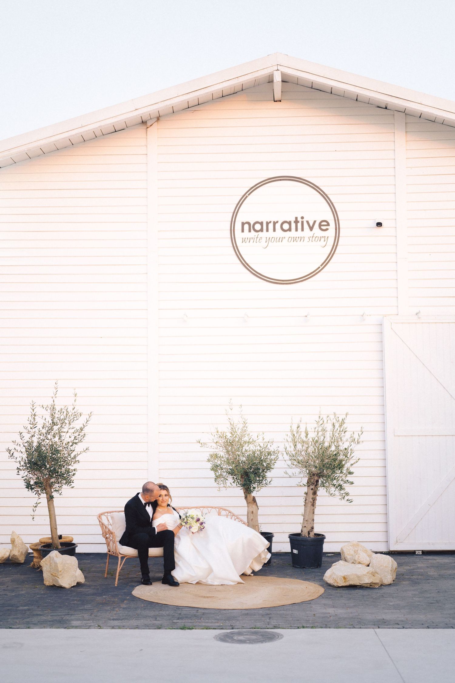 A romantic wedding scene with a couple seated on chairs against a white barn facade with olive trees and a 'narrative' logo.