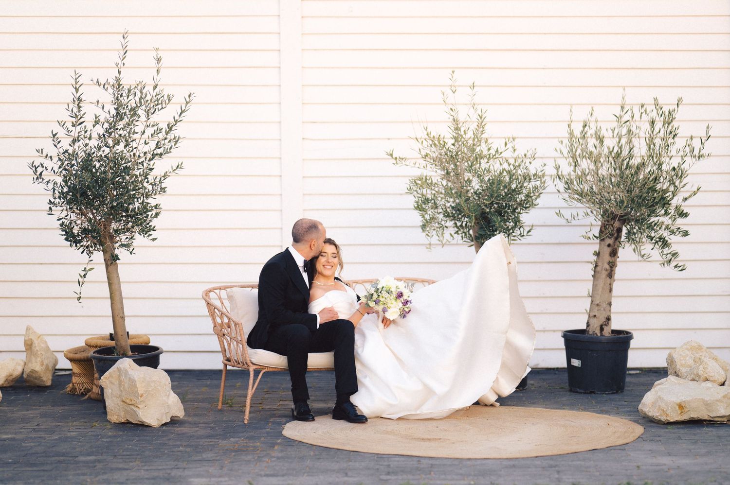 Elegant wedding couple sits together on outdoor patio with potted olive trees against white wall.