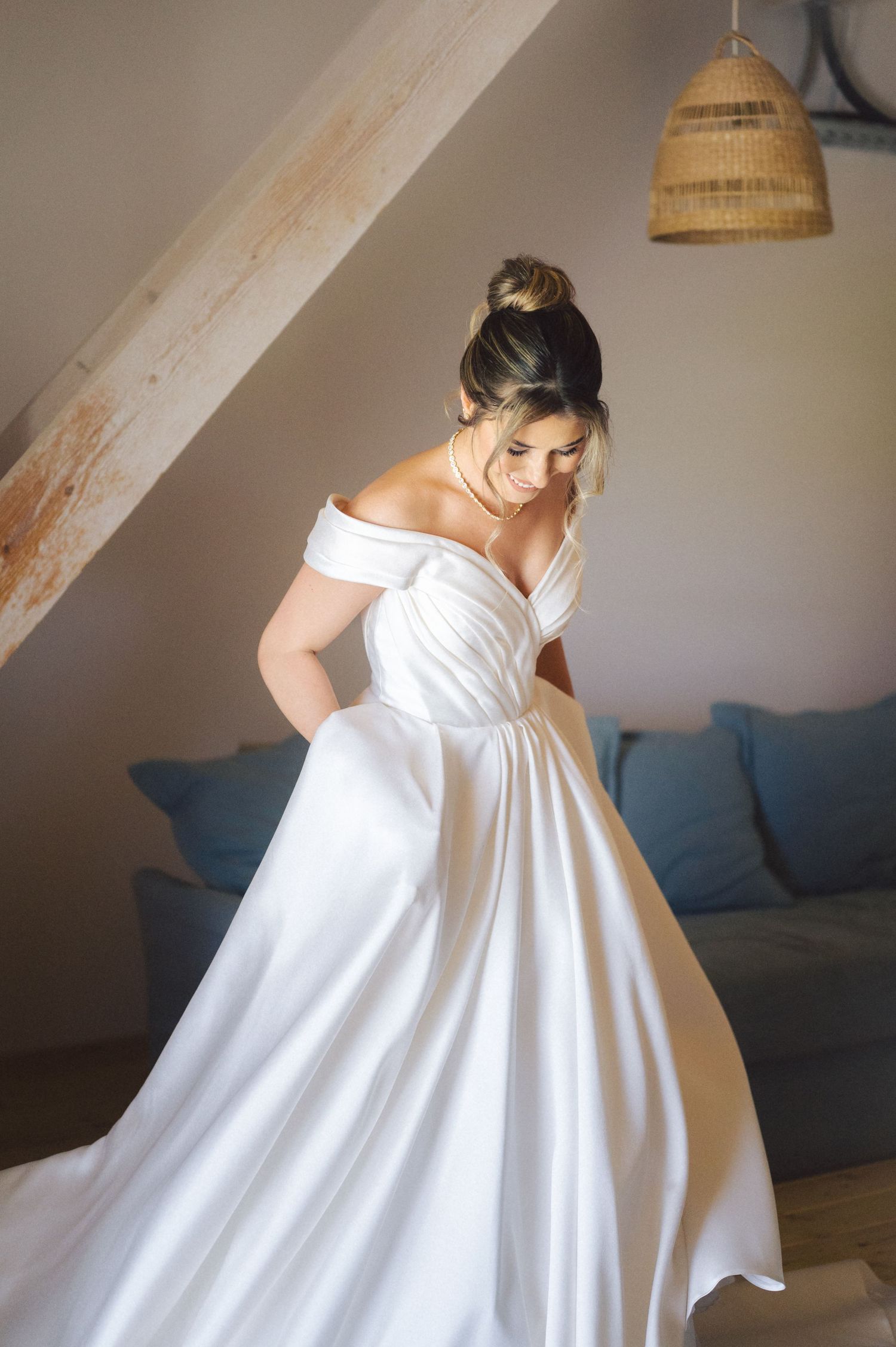Elegant off-shoulder white wedding gown with full skirt and natural lighting under rattan pendant lamp in cozy setting.