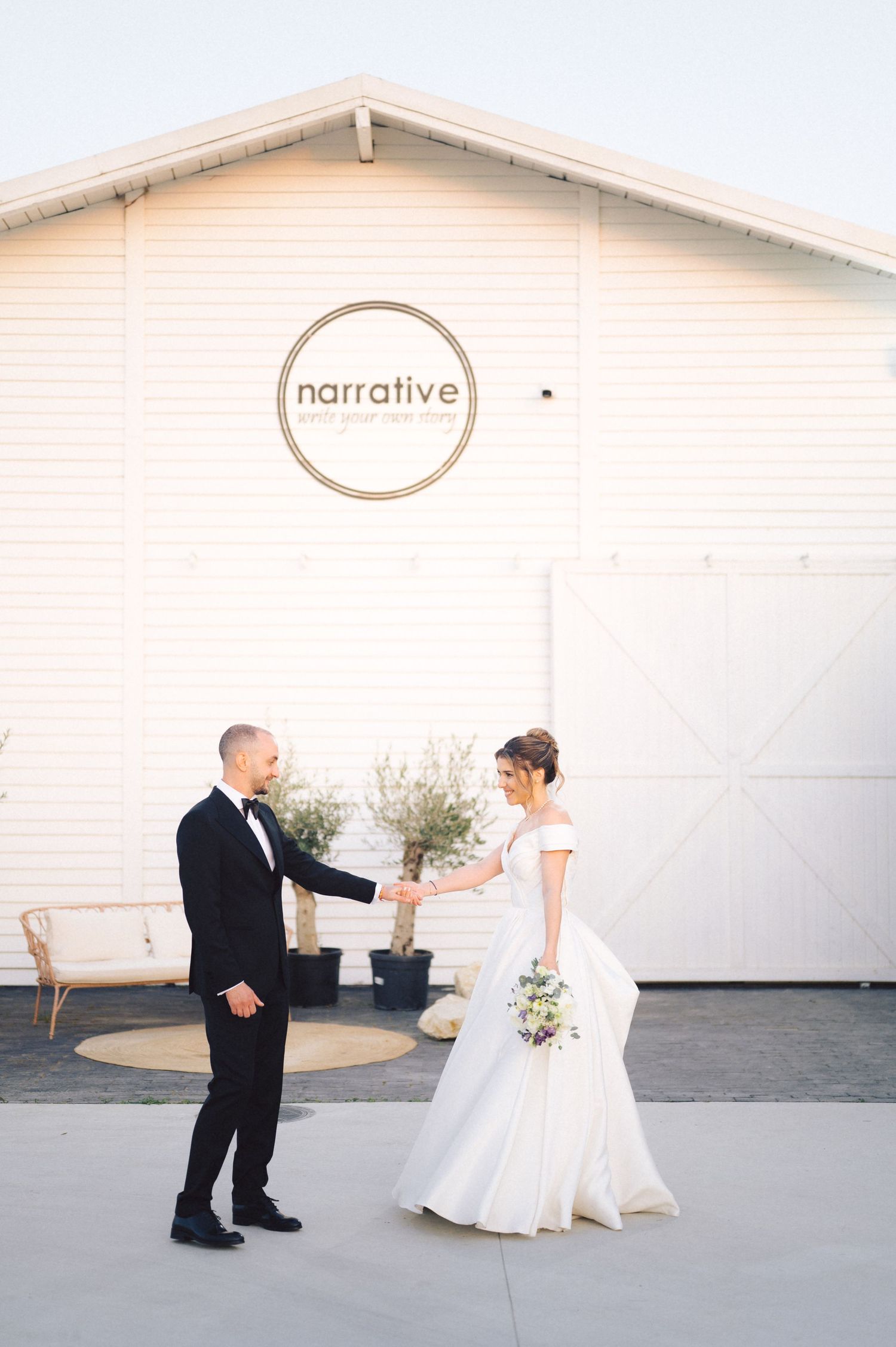 A couple shares a joyful first look moment outside a white building with a 'narrative' logo during their wedding celebration.