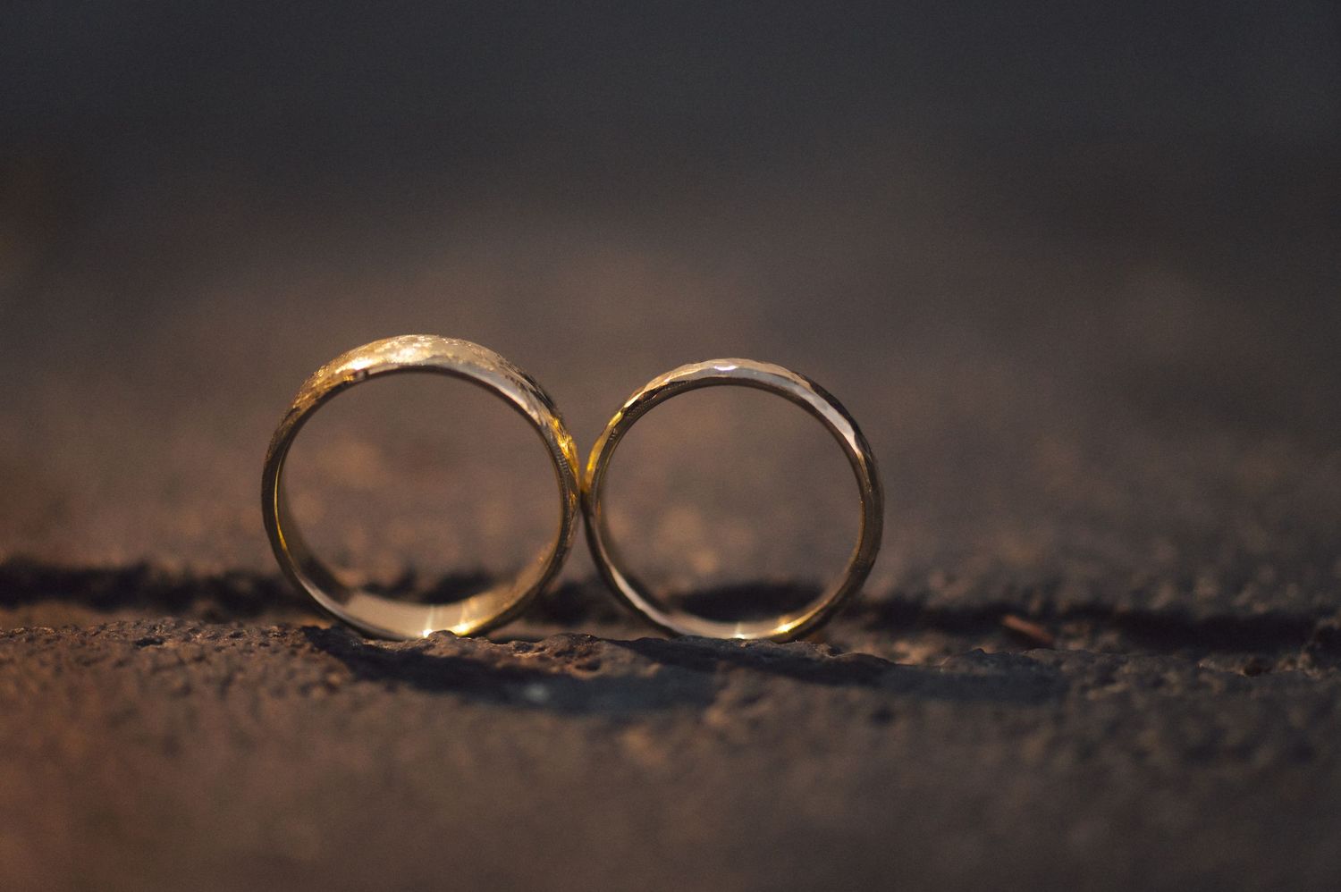 Two golden wedding rings lie close together on a textured brown surface in moody lighting.