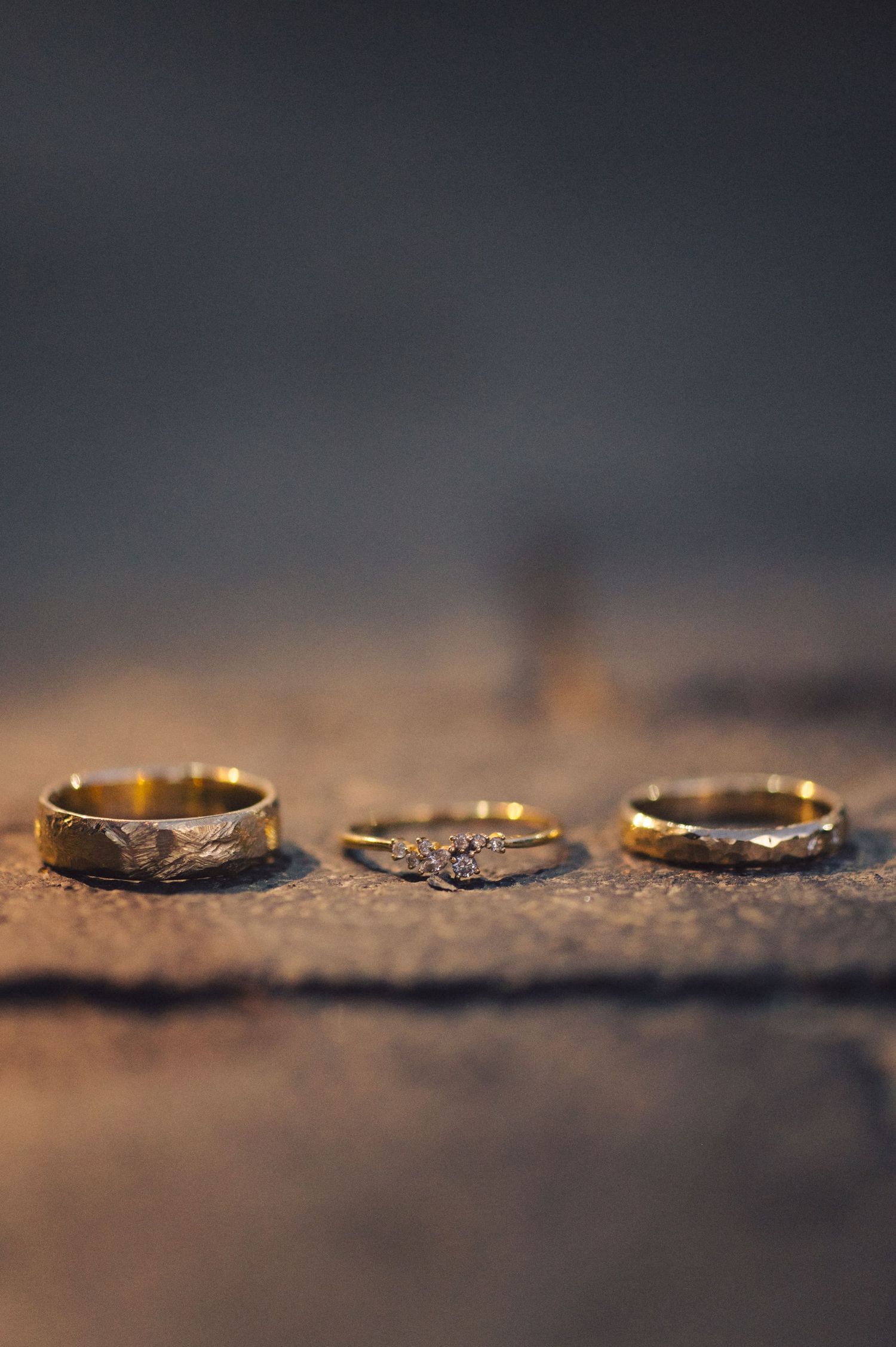 Three gold wedding rings with intricate designs arranged on a textured brown surface in soft lighting.