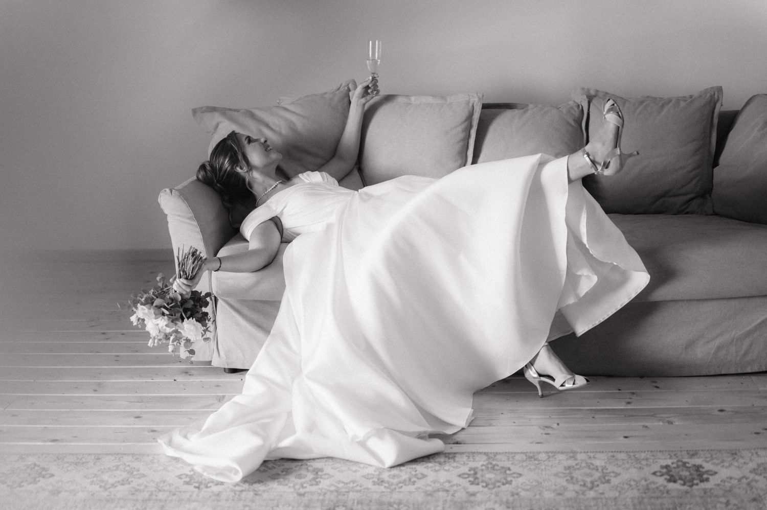 A figure in a flowing white dress reclines dramatically on a gray couch beside scattered flowers.