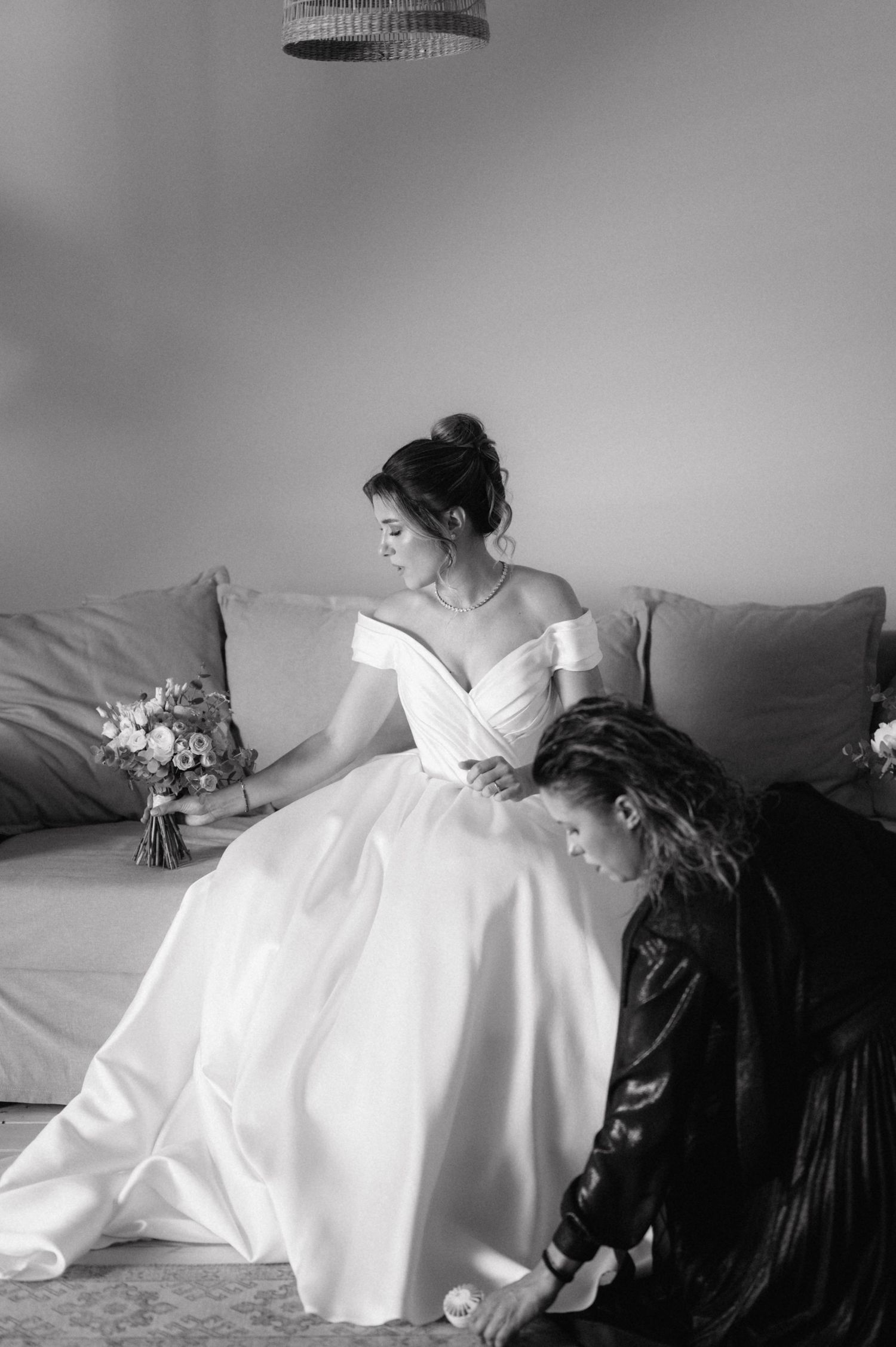 A black and white wedding photo showing a bride in an elegant off-shoulder gown being attended to on a couch.