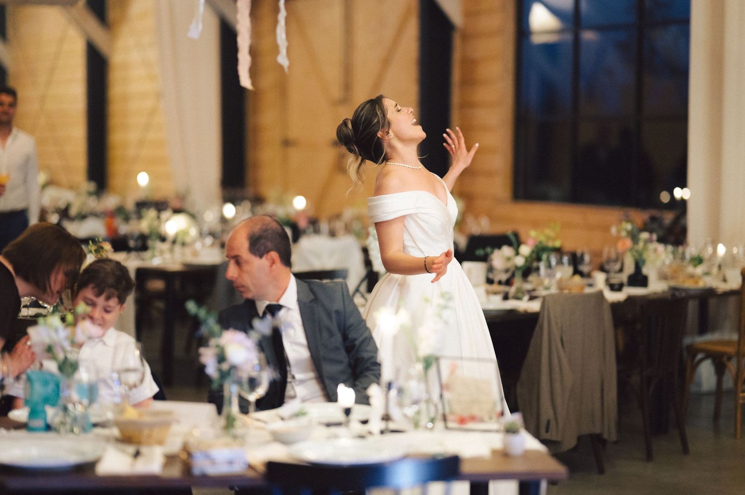 A lively wedding reception scene shows a person in a white dress joyfully dancing while others sit at elegantly set tables.