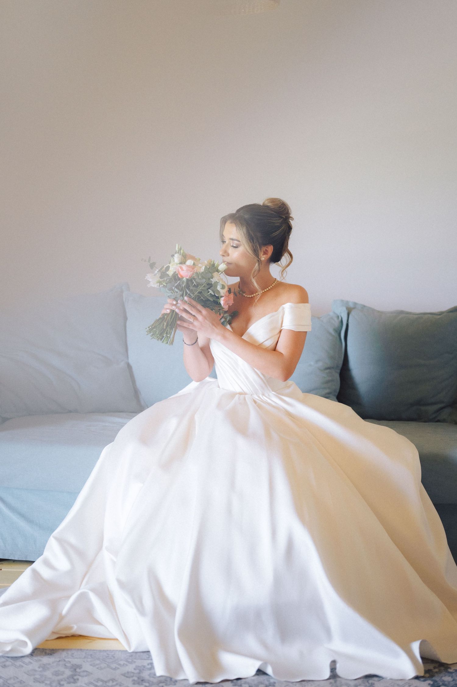 A bride in an off-shoulder white ballgown sits on a gray couch while smelling her pink and white bouquet.