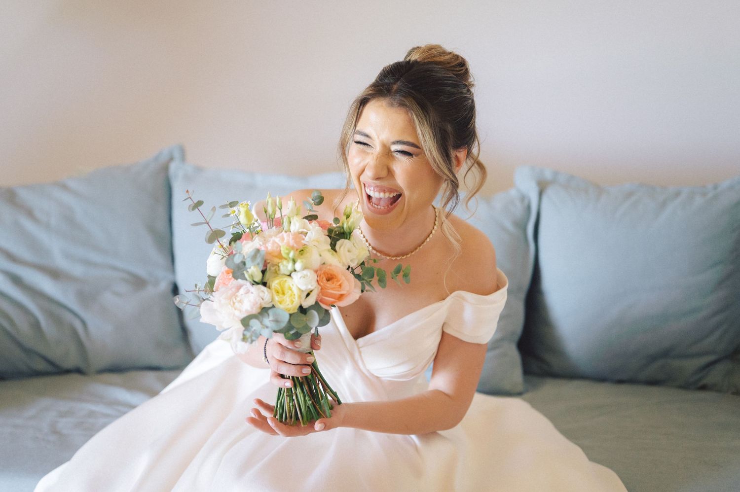 Joyful bride laughs while holding a pastel spring bouquet while seated on a light blue couch in a white wedding dress.