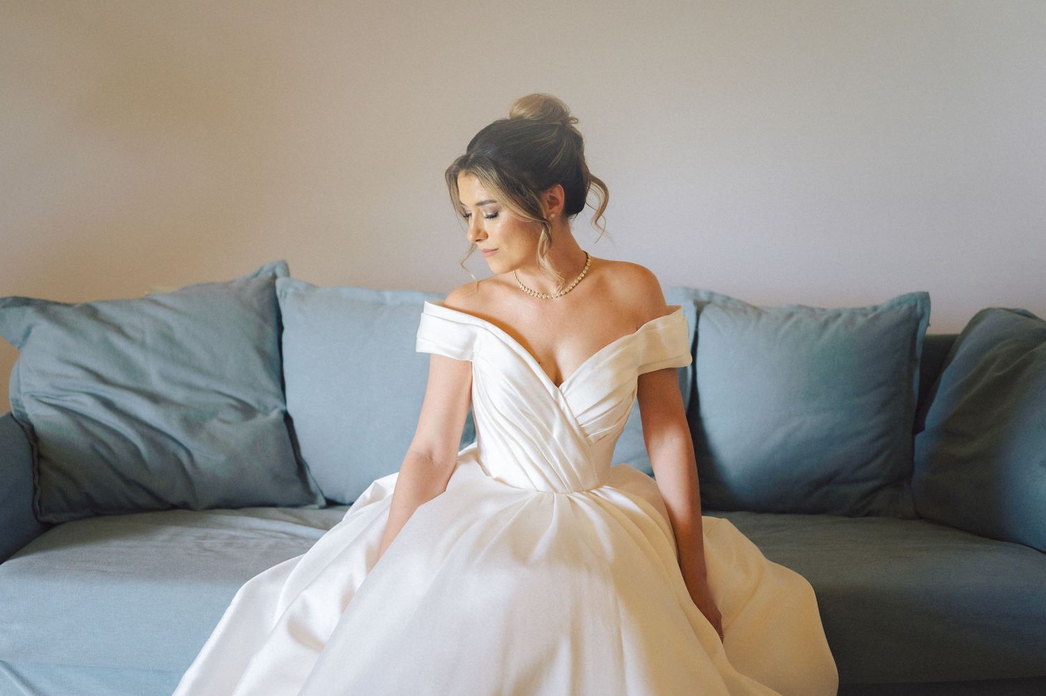Off-shoulder white wedding gown with gathered bodice and full skirt photographed against teal couch.