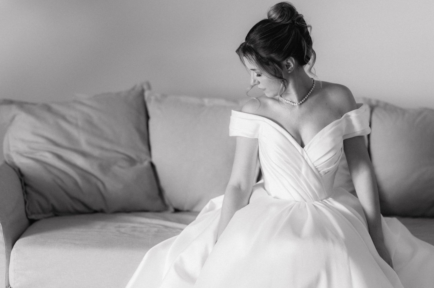 Black and white photo of a bride in an off-shoulder wedding gown sitting elegantly on a white couch.