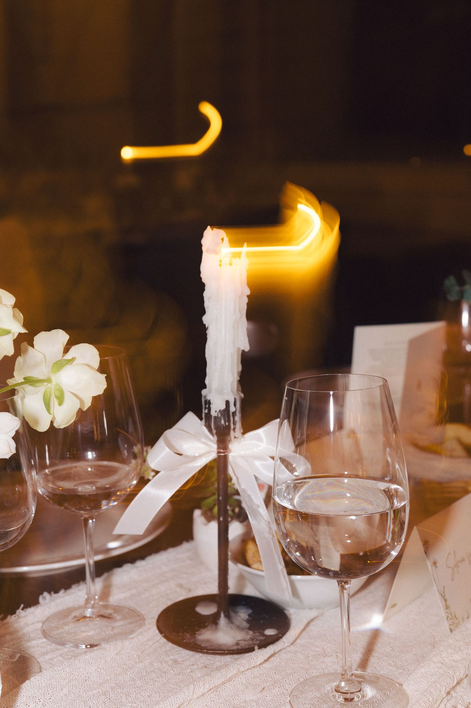 A candlelit table setting with wine glasses and white flowers on a tablecloth shows a melted candle dripping wax.