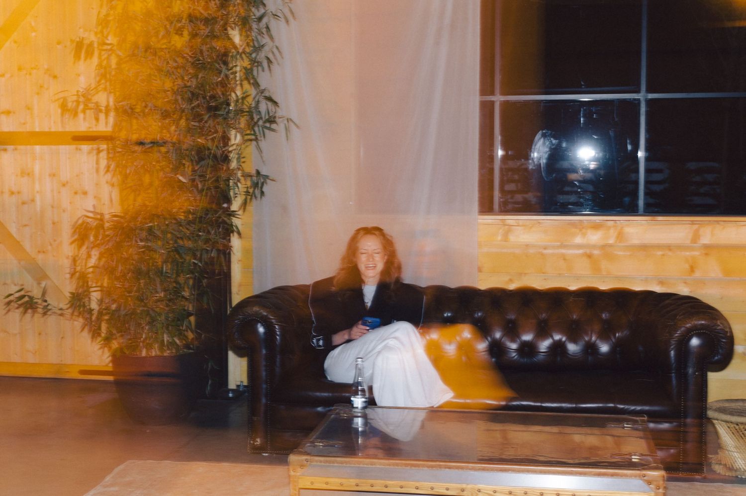 A person lounges casually on a black leather Chesterfield sofa in a warmly lit room with large windows.