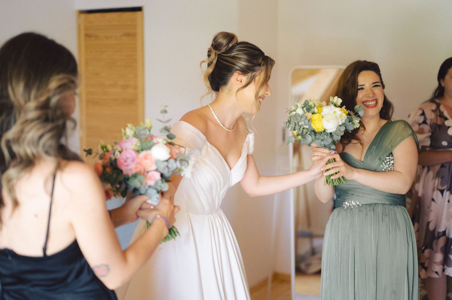 A bride in white dress surrounded by bridesmaids in sage green dresses share joyful moments with bouquets at wedding.