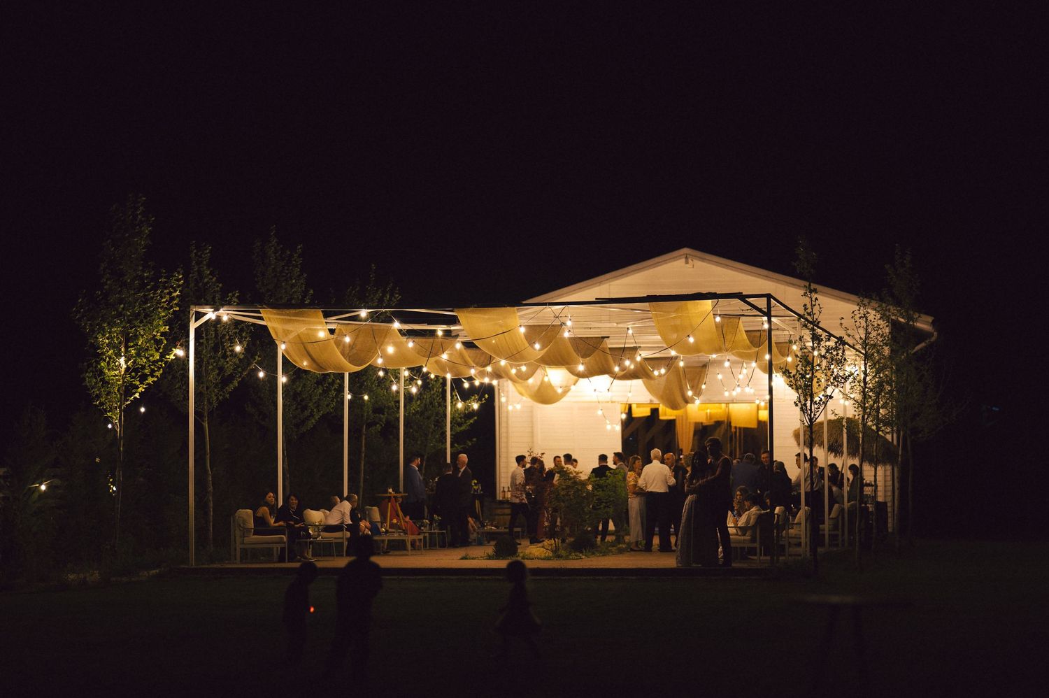 An outdoor wedding reception tent glows warmly at night with string lights and guests celebrating inside.