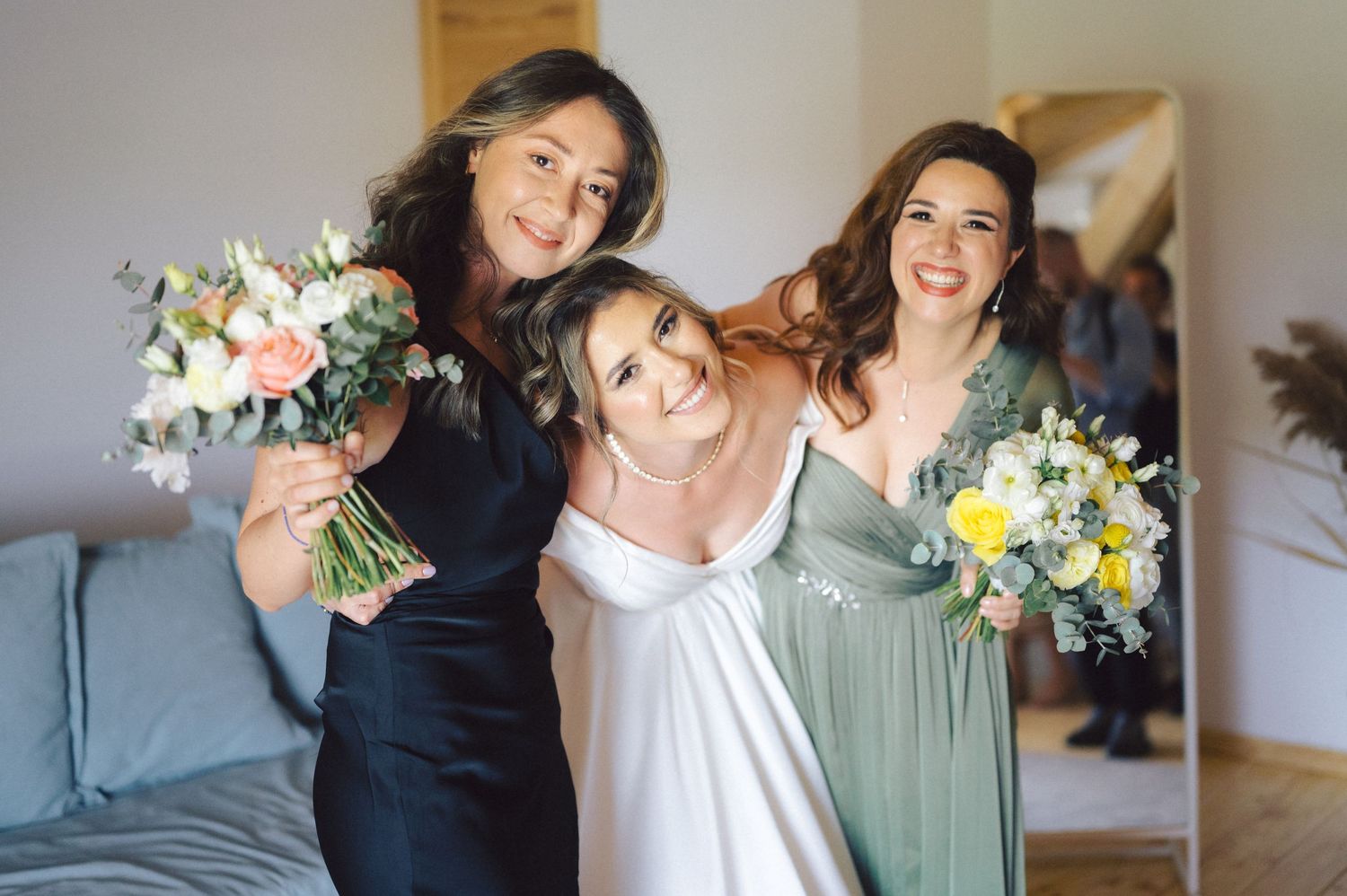 Three people in wedding attire pose together with bouquets of blush and white flowers while sharing joyful smiles.