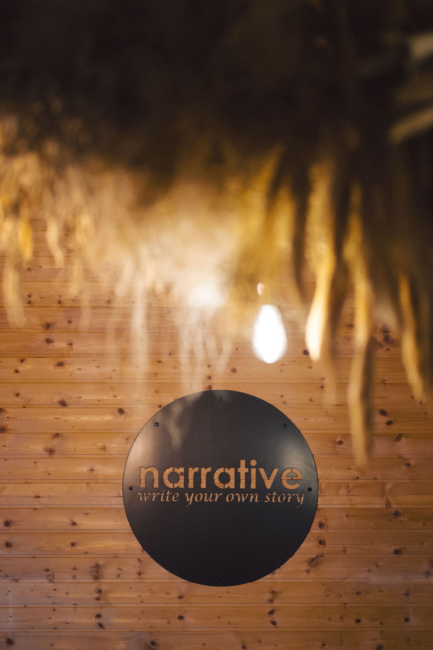 Illuminated wooden wall with black circular sign reading 'narrative - write your own story' in moody lighting.