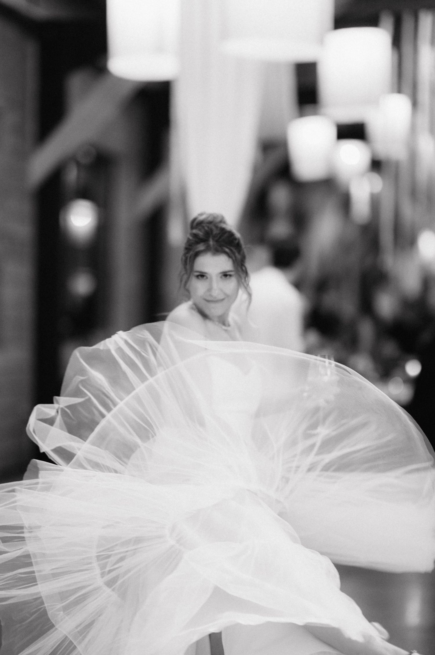 A black and white photograph showing a elegant bridal gown with flowing tulle fabric in an indoor setting with bokeh lighting.