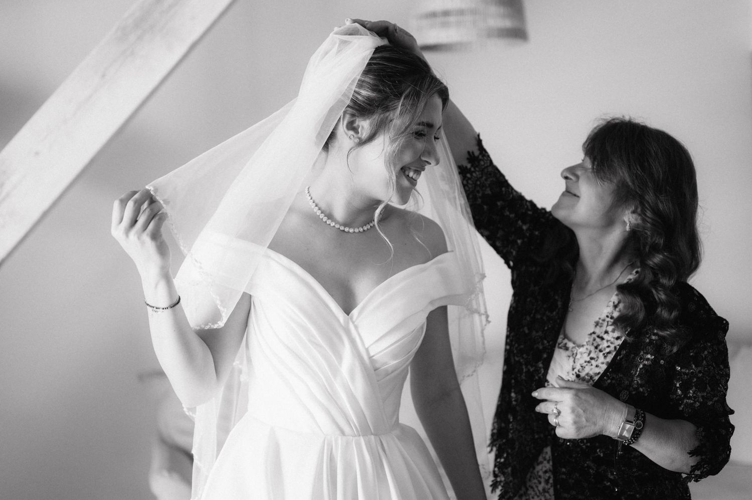 A tender moment of wedding preparation as a veil is adjusted on a bride wearing an elegant off-shoulder white gown.