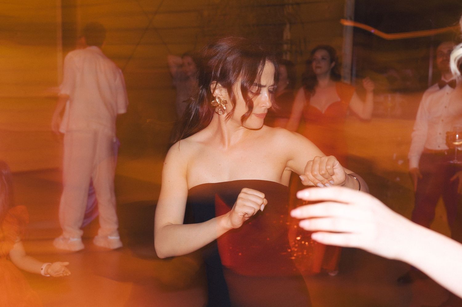 Person in black strapless dress reaching for a drink at a dimly lit party with an orange-tinted ambiance.
