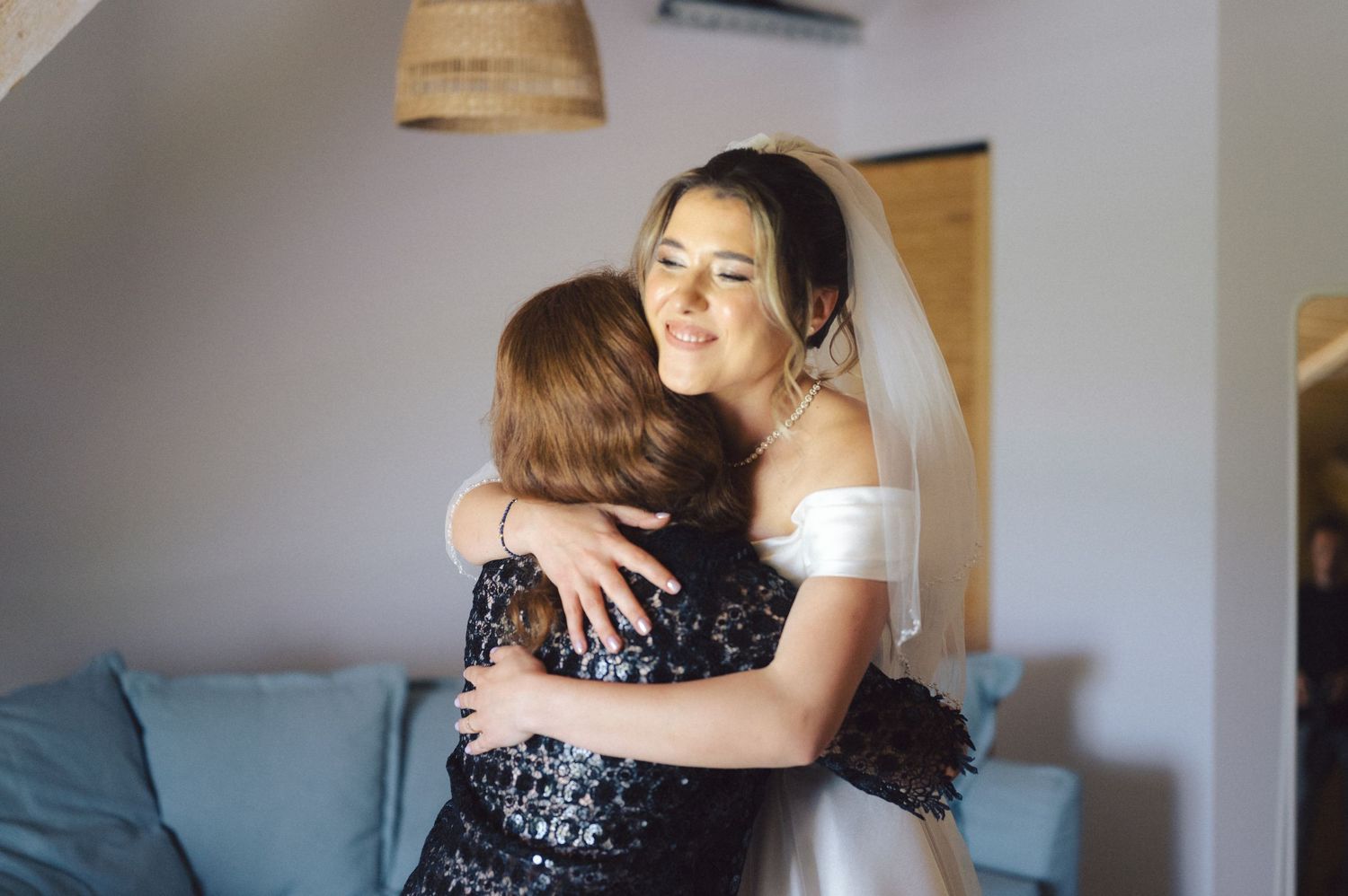 Two people share a tender embrace while sitting on a couch in a sunlit room with a woven lamp shade visible above.
