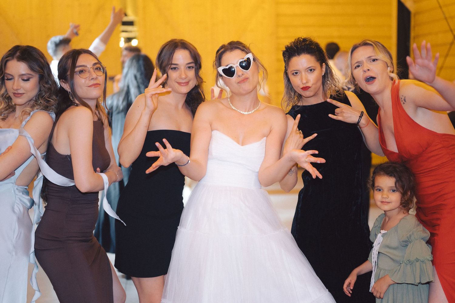 A group of women in formal attire strike playful poses together at a wedding celebration against a yellow backdrop.