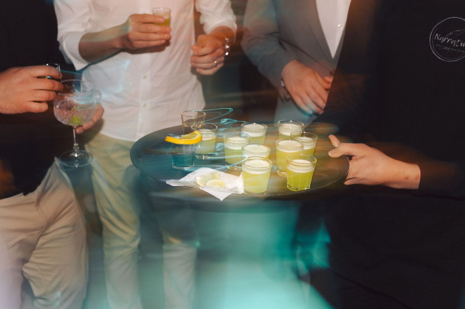 People holding yellow cocktail drinks on tray at party with blurred motion effect in dark setting.