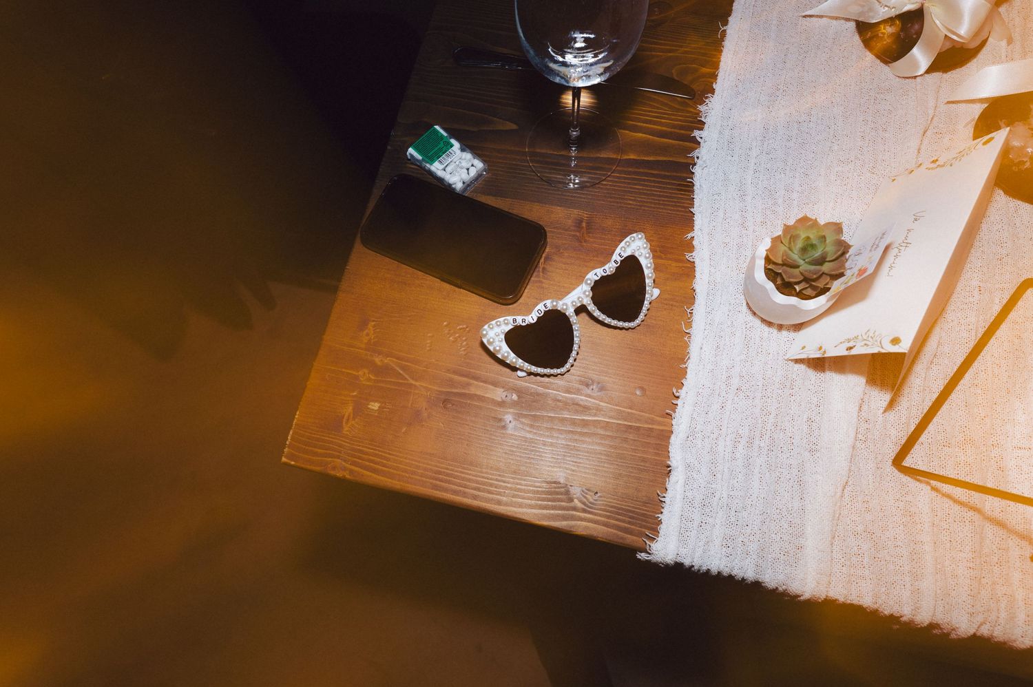 White heart-shaped sunglasses and wine glass on wooden table with white tablecloth in warm lighting.