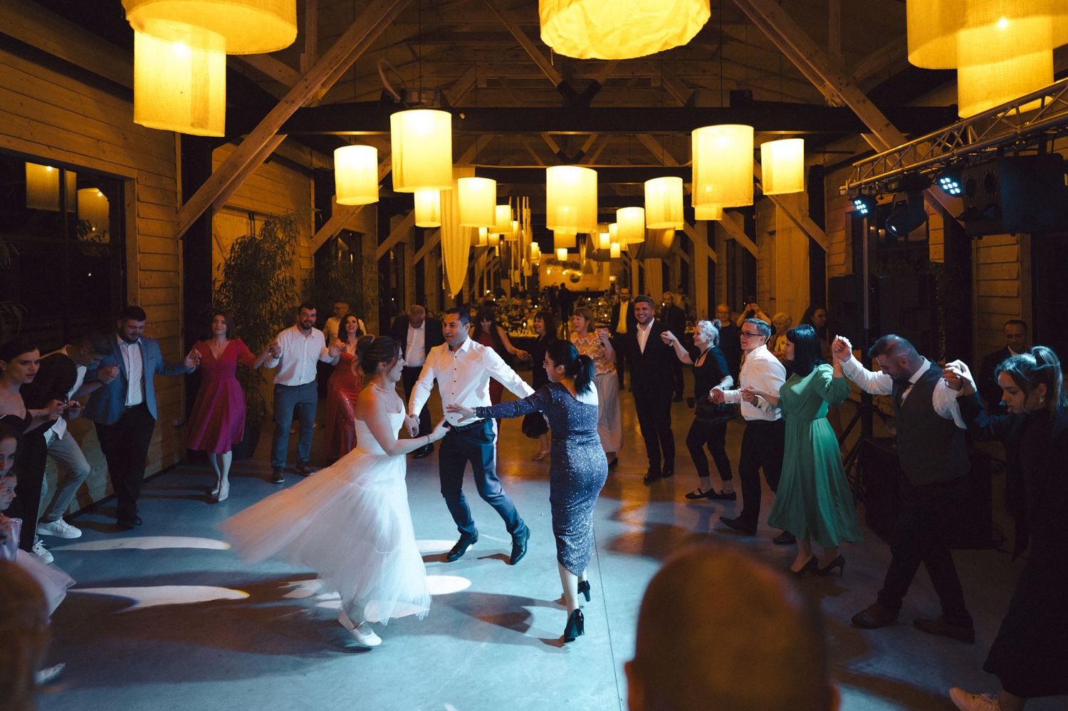 A lively wedding reception dance floor illuminated by warm glowing lanterns as guests celebrate under wooden beams.