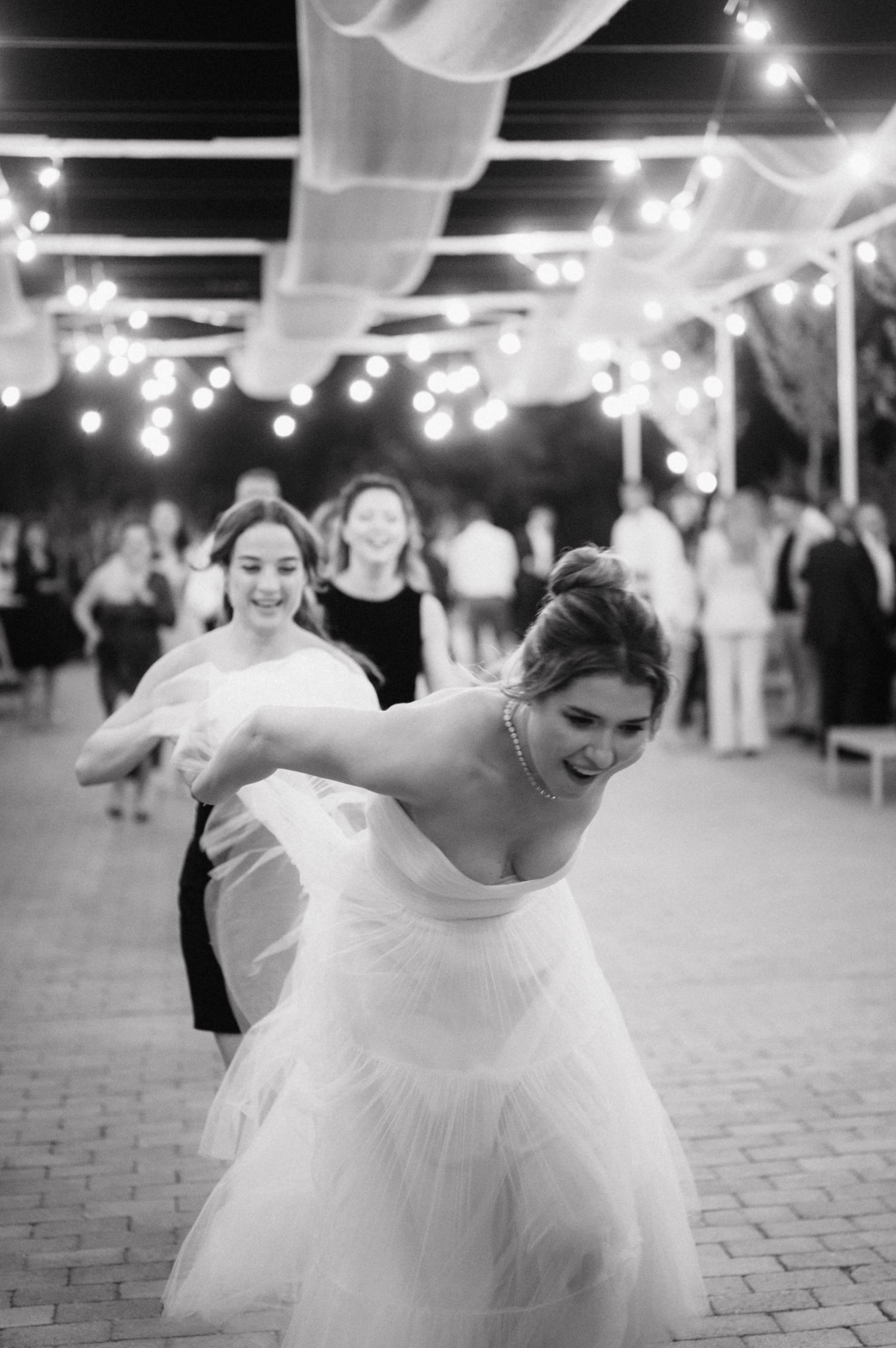 People dance and celebrate at an evening wedding reception under draped fabric and string lights on a brick patio.