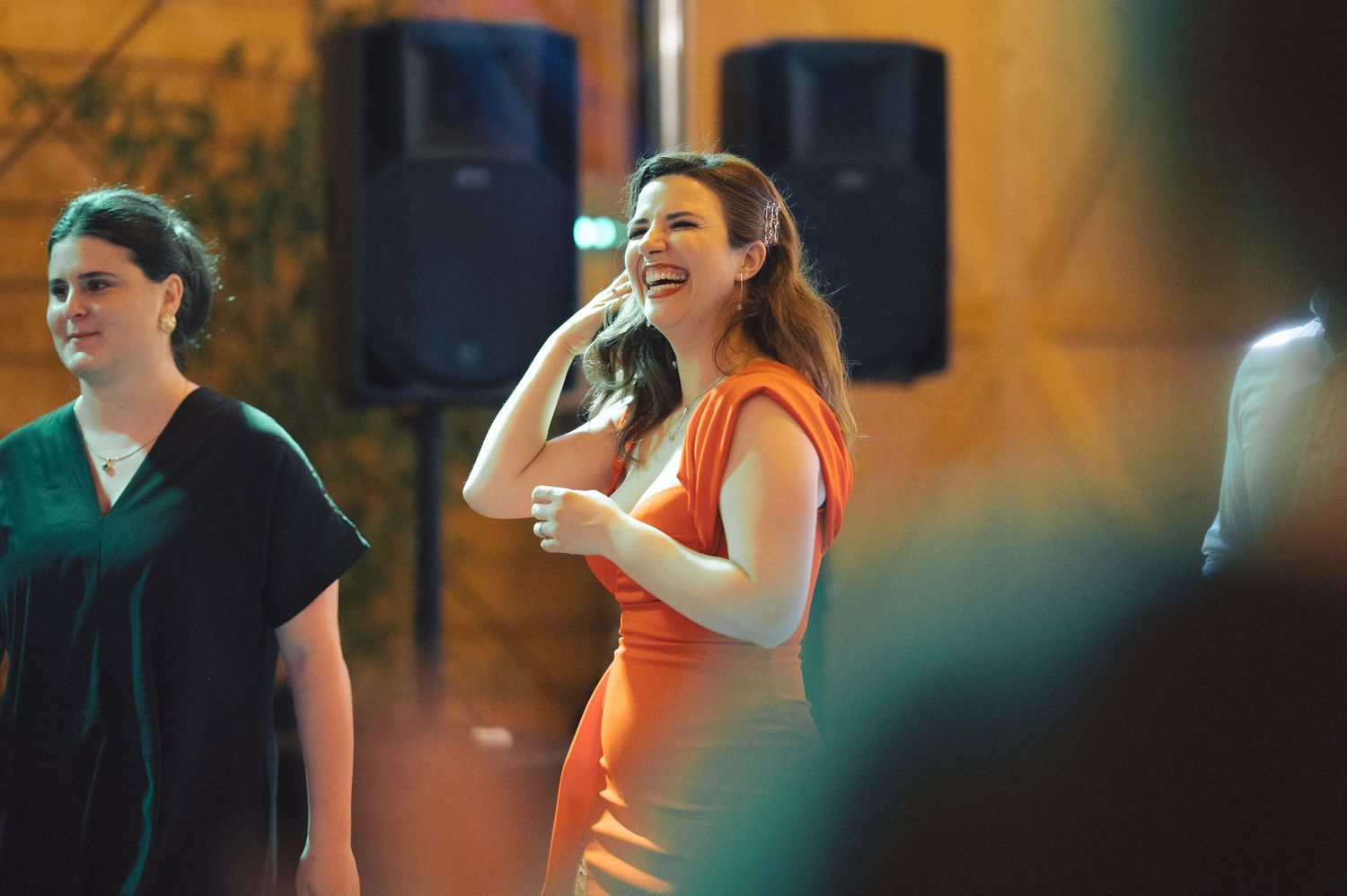 A person in a fitted orange dress laughs joyfully in front of speaker equipment during an indoor event.