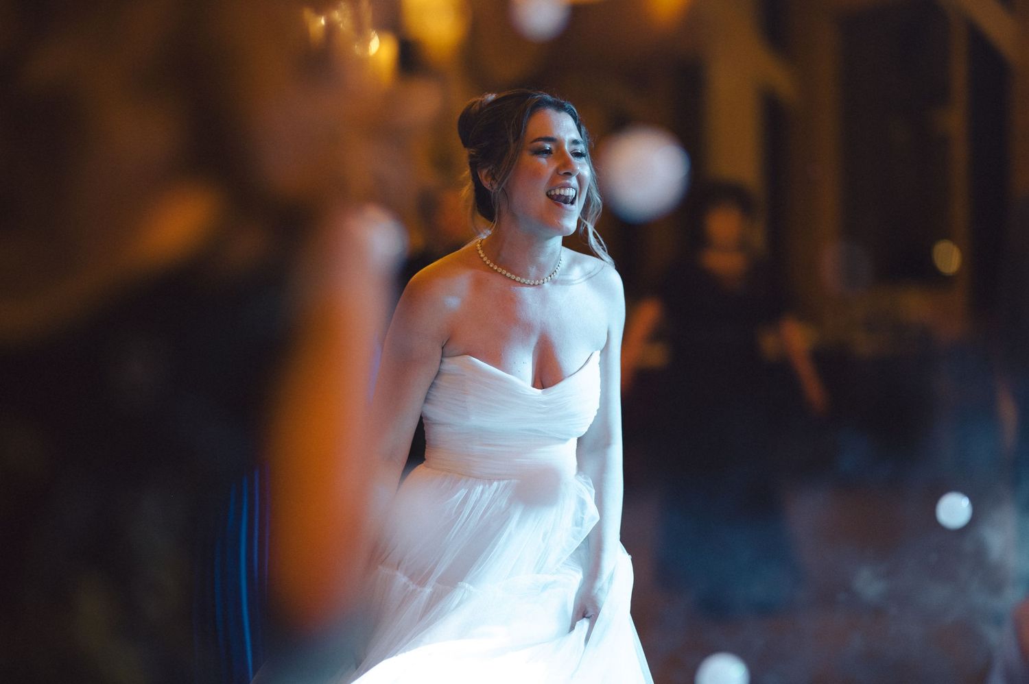 A bride in a white strapless wedding gown laughs joyfully during an evening celebration with bokeh lights in the background.