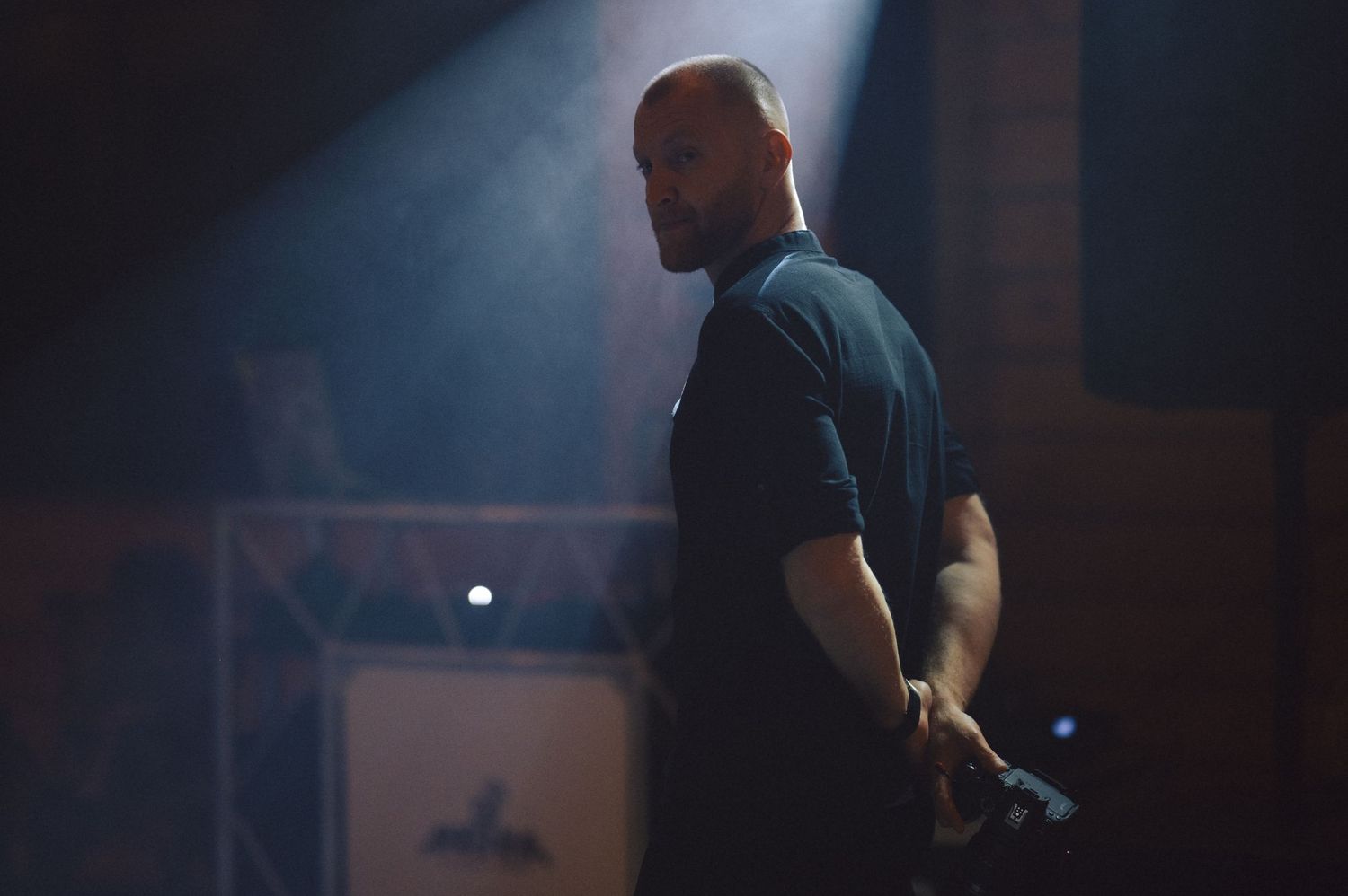 A person in a dark shirt stands in dramatic backlighting with smoke effects on stage.